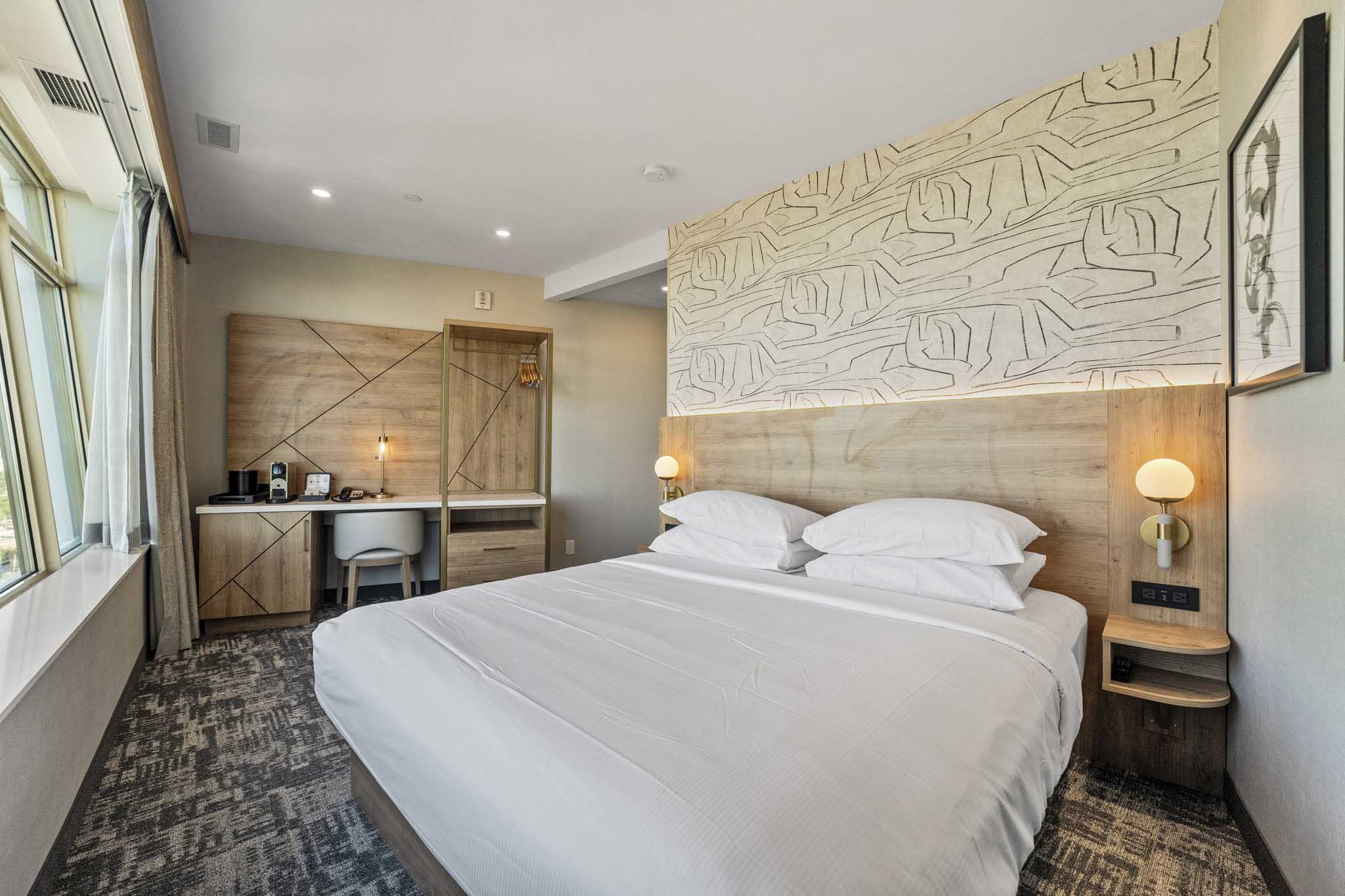 A hotel room overlooking the city with a king bed with white linen and a spacious desk for work or study purposes.