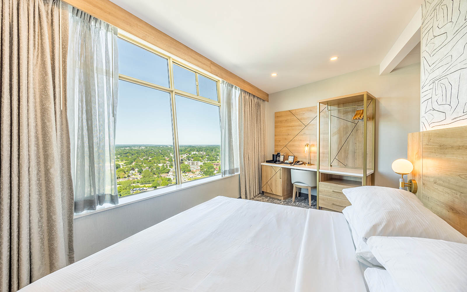 A hotel room overlooking the city with a king bed with white linen and a spacious desk for work or study purposes.
